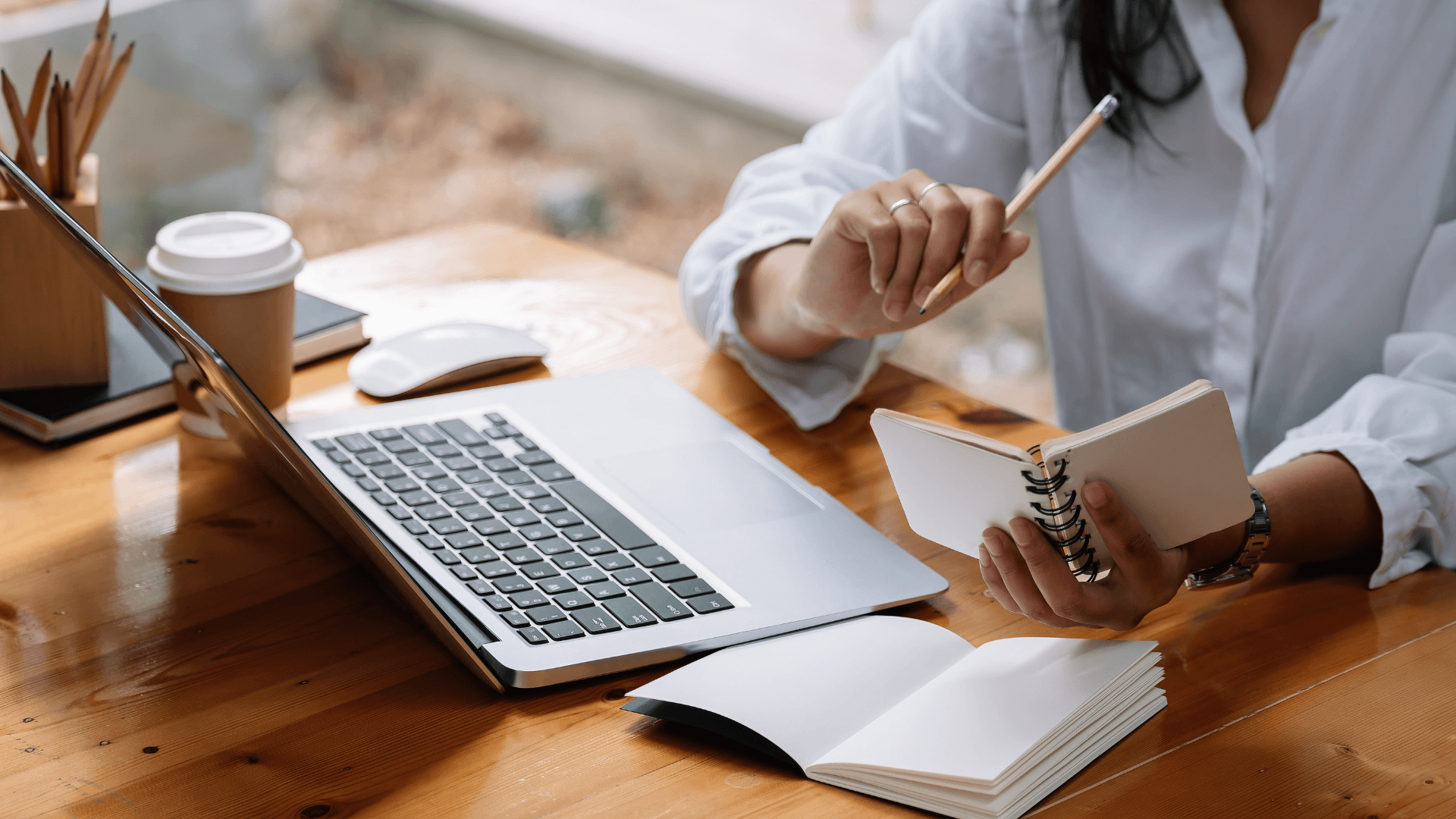 Person taking notes during online training session, with a laptop, notebook, and coffee on a desk.